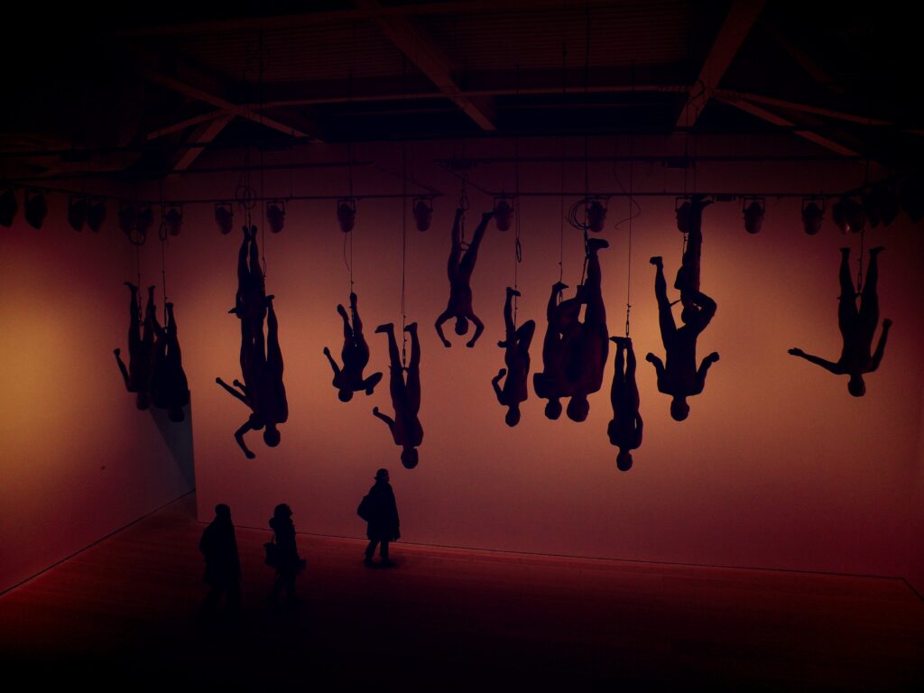 ein riesiger dunkler Ausstellungsraum, in dem an den Füßen aufgehängte Figuren von der Decke baumeln und einige Besucher zu ihnen hochschauen. Der Raum ist in fahles rötliches Licht getränkt.