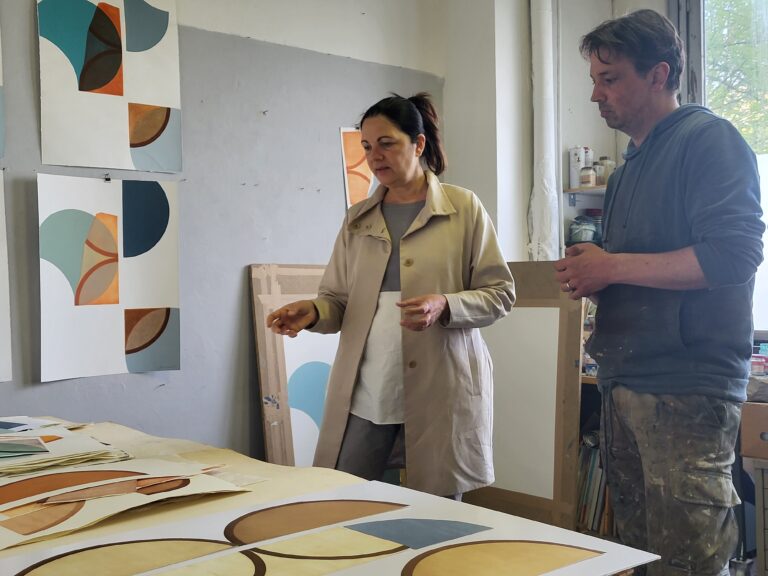 Die Kuratorin Dr. Margit im Schlaa spricht über die Kunstwerke des Künstlers Pedro Boese in dessen Atelier. Der Künstler hört aufmerksam zu.