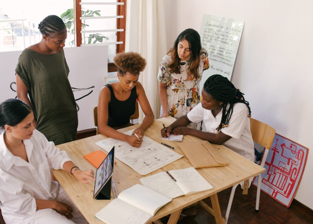 Fünf junge Frauen verschiedener Nationalitäten sitzen bzw. stehen an einem Tisch herum, an dem sie im Rahmen eines Gruppencoachings arbeiten.
