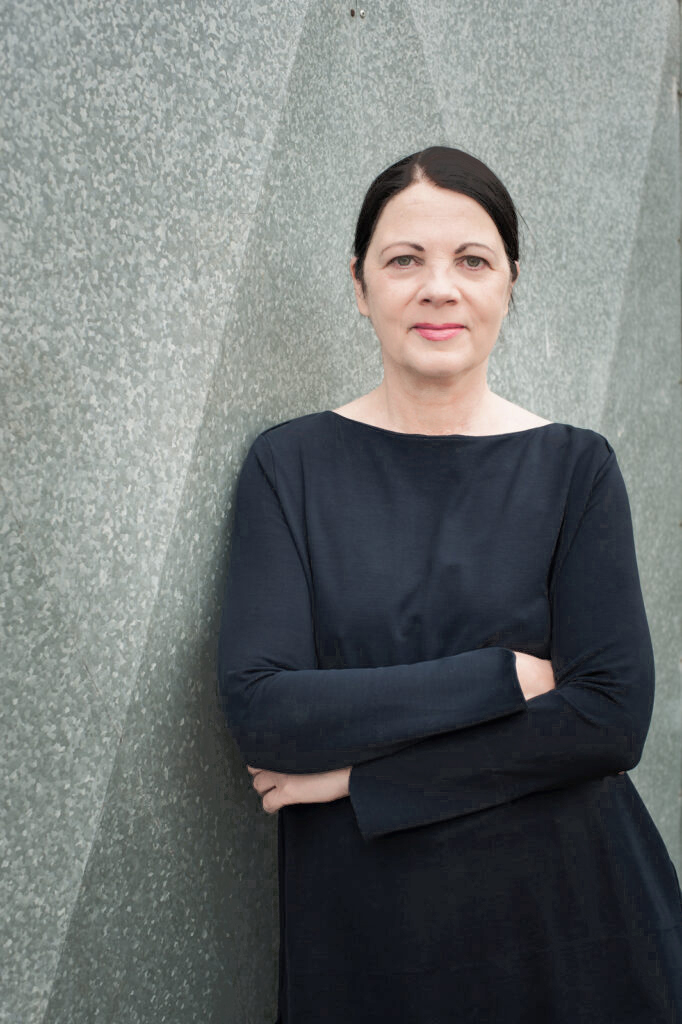 Ganzkörperporträt der Kuratorin Dr. Margit im Schlaa, die lächelnd mit verschränkten Armen den Betrachter anschaut.